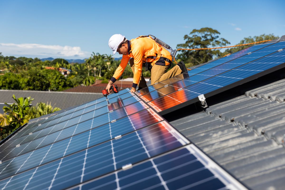 A technician in safety gear installs solar panels on a roof, surrounded by lush greenery and a clear blue sky.