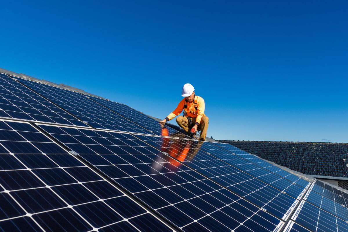 A worker in safety gear inspects solar panels on a roof under a clear blue sky. The panels reflect sunlight, showcasing their efficiency.