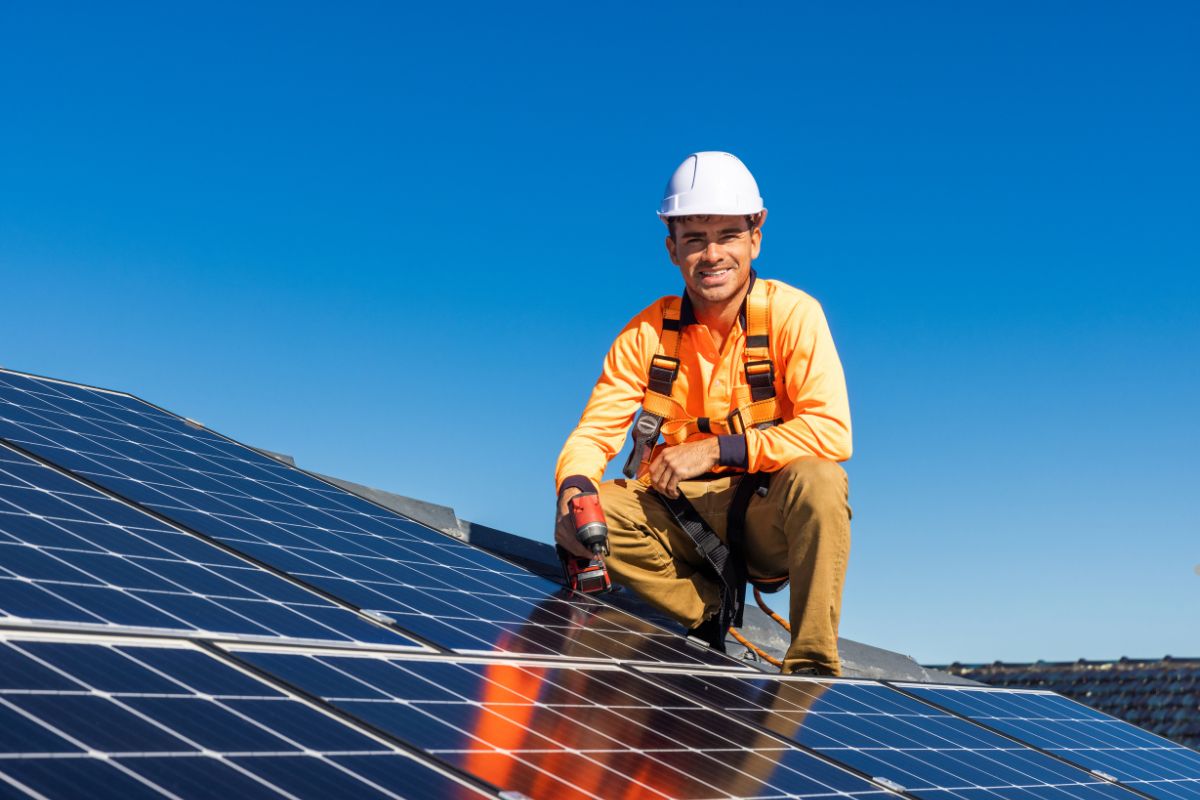 A worker in a safety harness and bright orange shirt installs solar panels on a rooftop under a clear blue sky.