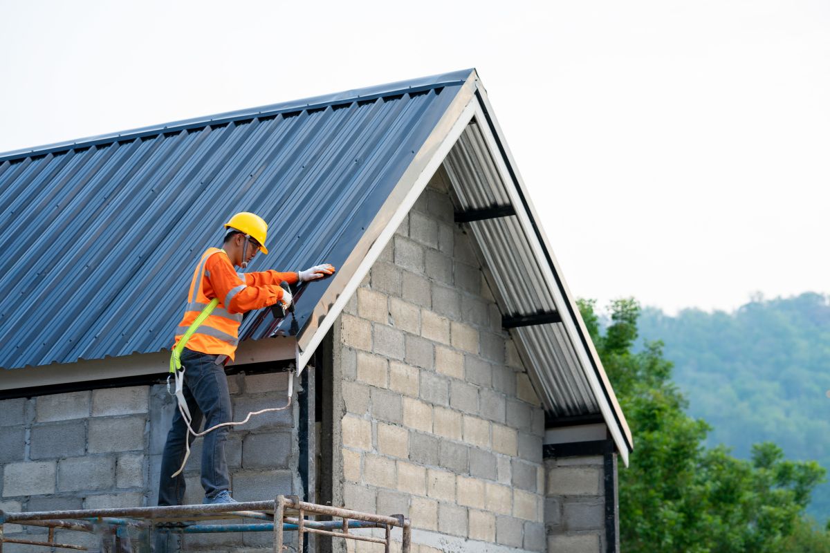  man in an orange vest and hard hat installs a drip edge on a house roof, focused on his work.