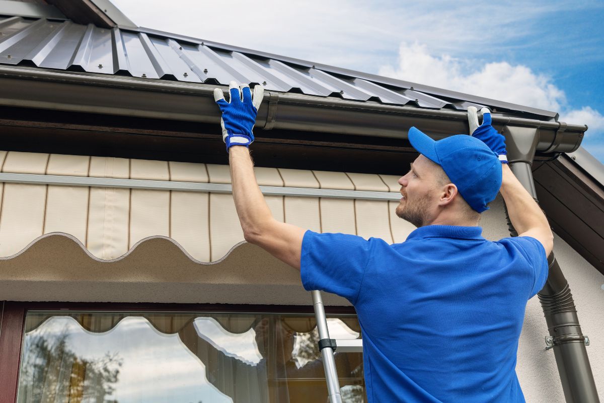 A man in a blue shirt and gloves installs a drip edge on a metal roof, focusing on precise adjustments. 